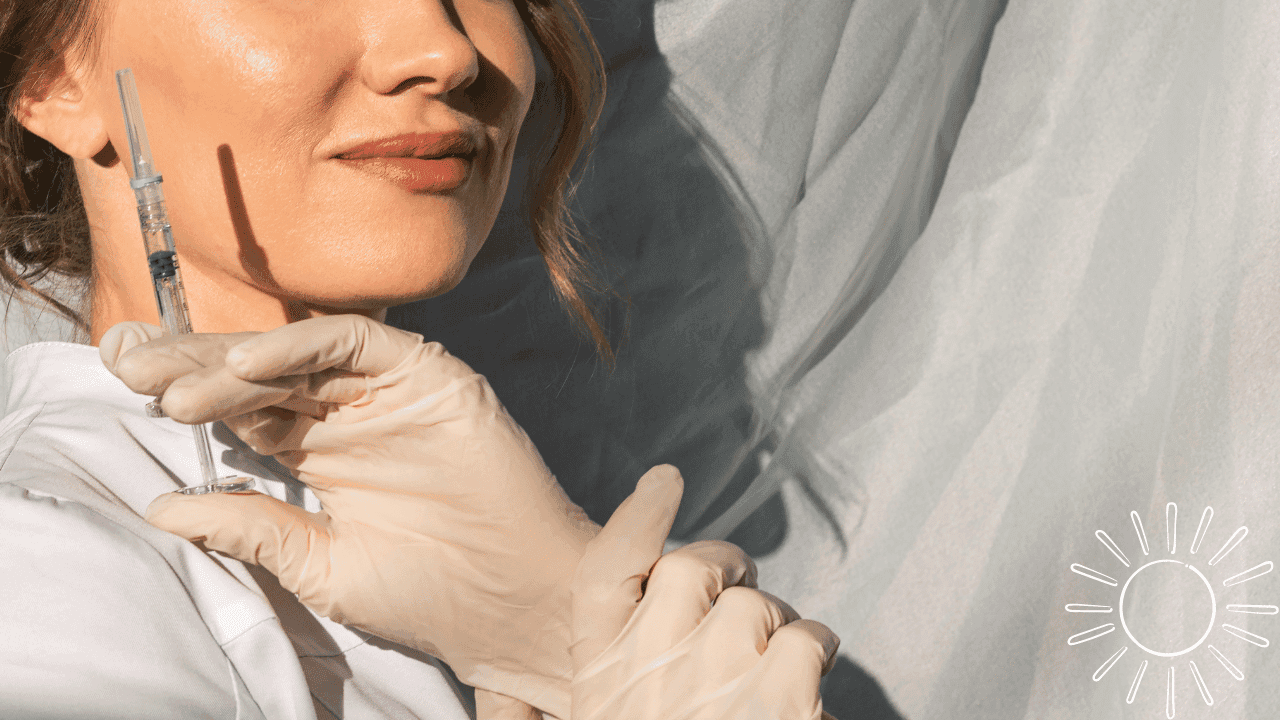 Detailní záběr na ženu v bílém plášti, která má na rukou latexové rukavice a drží injekční stříkačku. Její výraz působí profesionálně a klidně, zatímco její tvář i bílé pozadí, před kterým stojí, osvětluje sluneční světlo.
