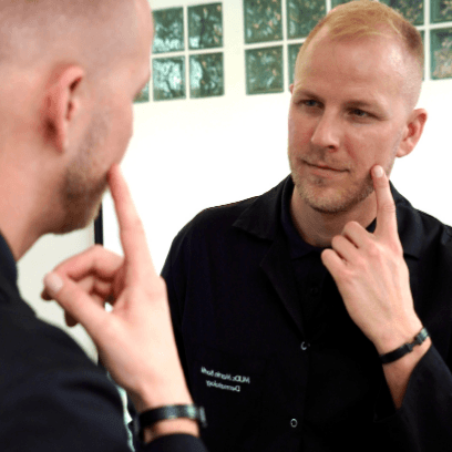 Dermatolog MUDr. Martin Bortlík ze Skinity Clinic se dívá do zrcadla a ukazuje si na tvář.